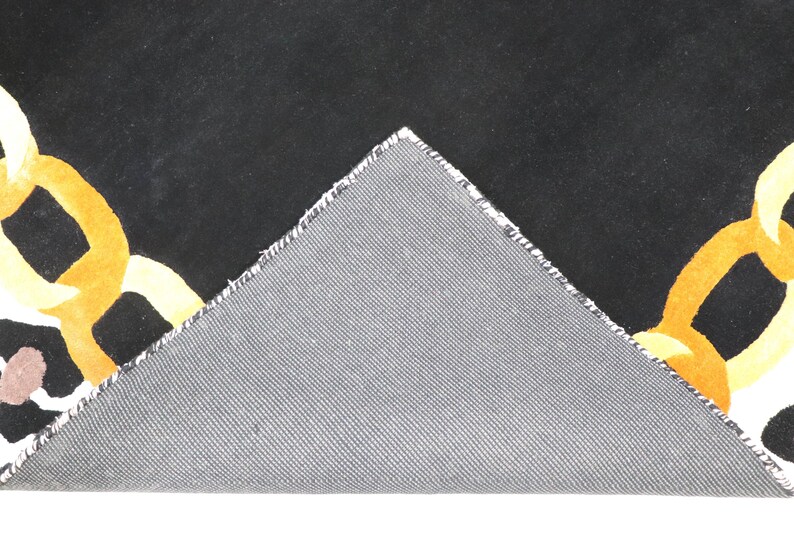 May include: A black rug with a yellow and orange chain link pattern, featuring a triangular corner folded over to reveal a grey, textured, woven backing material. The rug displays a bold graphic design with interlocking links on a dark field.