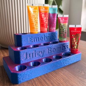 Könnte beinhalten: Ein blau-violetter, 3D-gedruckter, abgestufter Präsentationsständer mit der Gravur Emely's Juicy Bombs auf der Vorderseite. Der Ständer enthält fünf kleine Tuben Essence Juicy Bomb Lipgloss in den Farben Orange, Rot, Pink und Grün.