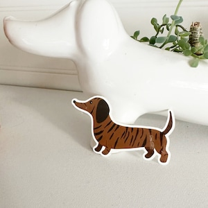 May include: A brown and black dachshund sticker with a white background. The dachshund is illustrated in a cartoon style with a striped pattern.