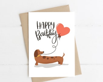 May include: A white card with the text "Happy Birthday" in black script. A brown dachshund with a yellow collar is holding a red heart-shaped balloon on a string.