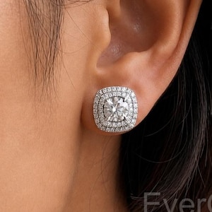 May include: A silver-toned stud earring featuring a round centre stone surrounded by a double halo of smaller pavé-set stones in a square-shaped frame. The earring is worn on an earlobe. The text EverGlowJewels is visible in the bottom right corner.