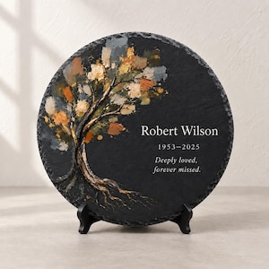 Puede incluir: Una placa conmemorativa circular de pizarra negra con un árbol pintado de hojas texturizadas doradas, marrones y grises. El texto dice Robert Wilson, 1953-2025, y Deeply loved, forever missed. La placa se apoya en un soporte de metal negro.