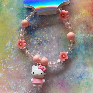 May include: A beaded bracelet featuring clear faceted beads, matte pink spherical beads, and pink flower-shaped beads with yellow centres. A white cat character charm with a pink bow and pink dress hangs from the centre of the elasticised band.