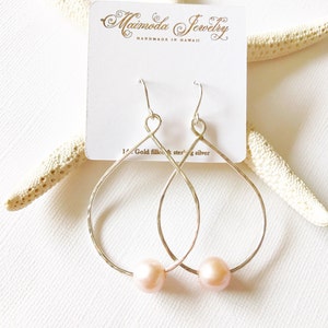May include: A pair of silver teardrop-shaped earrings with a delicate hammered texture. Each earring features a small pink pearl hanging from the bottom.