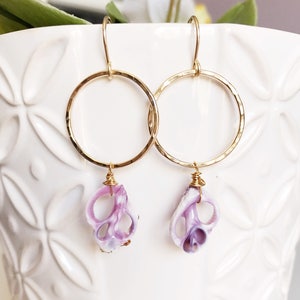May include: Gold hoop earrings with a hammered finish and a purple shell charm hanging from each hoop.