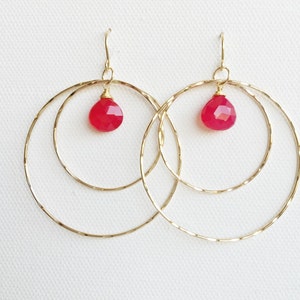 May include: Gold hoop earrings with a single red gemstone hanging from each hoop.