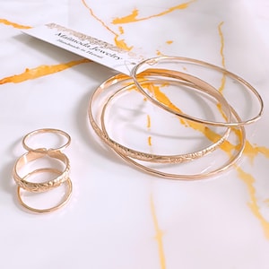 Op de afbeelding: Set van vijf gouden ringen met een bloemenmotief. De ringen zijn op elkaar gestapeld. De ringen liggen op een wit oppervlak met gele marmering. De afbeelding bevat ook een klein wit kaartje met de tekst "Mainoda Jewelry Handmade in Hawaii".