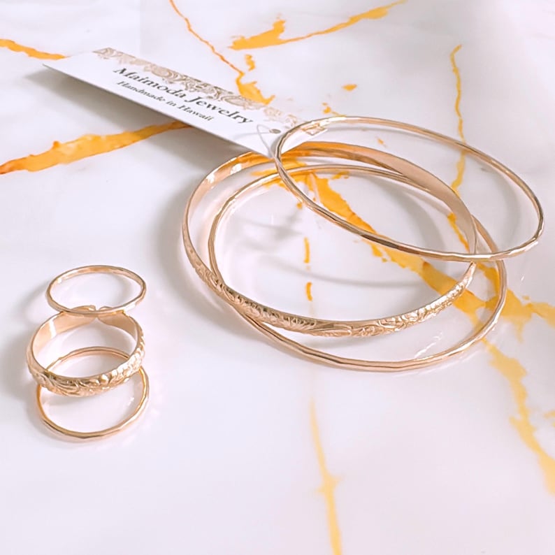 Op de afbeelding: Set van vijf gouden ringen met een bloemenmotief. De ringen zijn op elkaar gestapeld. De ringen liggen op een wit oppervlak met gele marmering. De afbeelding bevat ook een klein wit kaartje met de tekst "Mainoda Jewelry Handmade in Hawaii".