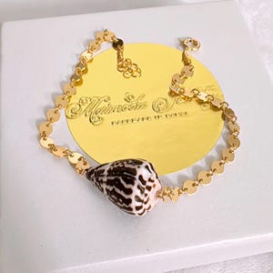 May include: Gold-toned bracelet featuring a natural seashell pendant with brown and white markings. The bracelet has a delicate chain design with small, round links. The seashell is the focal point, adding a touch of nature-inspired elegance.
