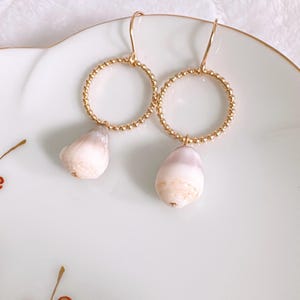 May include: A pair of gold-toned earrings featuring a textured, beaded circular hoop design. Each hoop suspends a smooth, pale pinkish-white teardrop-shaped shell charm. The earrings are attached to simple gold-toned ear wires for hanging.