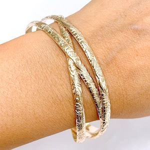 May include: Three gold bangle bracelets with intricate floral designs. The bracelets are stacked on top of each other and are worn on a person's wrist.