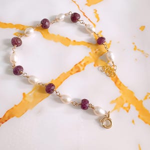May include: A delicate bracelet featuring white pearls and faceted, deep red gemstones strung on a gold-colored chain. The bracelet has a gold clasp and is displayed on a white surface with gold accents.