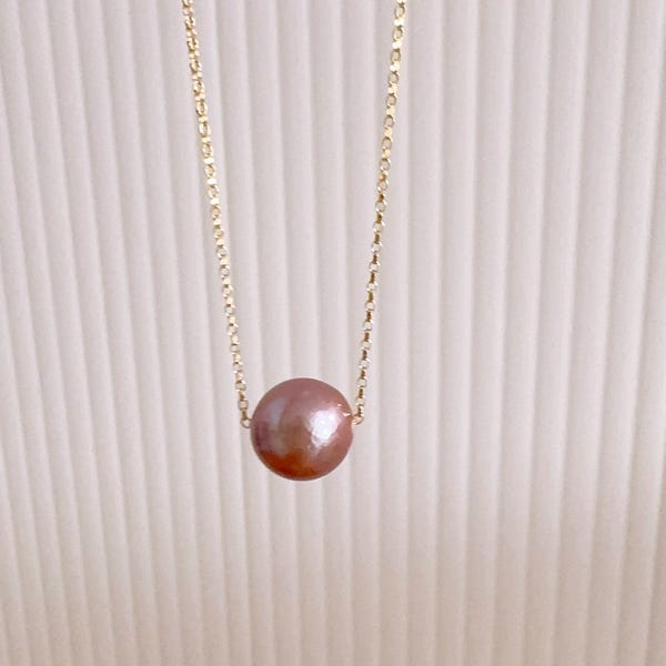 May include: A delicate gold chain necklace featuring a single, round, mauve-coloured pearl pendant centred on the chain. The necklace has a simple, refined design with a polished metallic finish on the chain and a smooth, lustrous surface on the pearl.