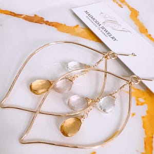 May include: Gold hoop earrings with a leaf design and dangling clear, pink, and yellow gemstones. The earrings are handmade in Hawaii by Maimoda Jewelry.