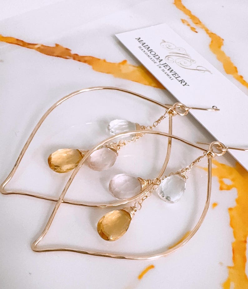 May include: Gold hoop earrings with a leaf design and dangling clear, pink, and yellow gemstones. The earrings are handmade in Hawaii by Maimoda Jewelry.