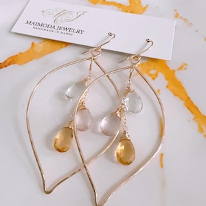 May include: Gold leaf earrings with a teardrop shape. The earrings feature three dangling stones: a light green, a light pink, and a light yellow.