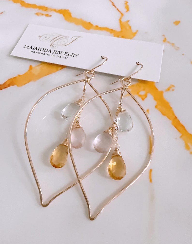 May include: Gold leaf earrings with a teardrop shape. The earrings feature three dangling stones: a light green, a light pink, and a light yellow.