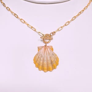 May include: Gold-toned necklace featuring a seashell pendant. The pendant is a light pink and yellow seashell with gold wire detailing. The necklace has a toggle clasp and a chain link design.