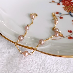 May include: A delicate gold-toned bracelet featuring a series of soft pink pearls. The bracelet is composed of a fine chain with evenly spaced pearls, creating an elegant and understated design. The bracelet is displayed on a white surface.