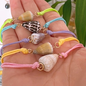 May include: A collection of seashell bracelets with colorful cords in neon green, blue, purple, yellow, and pink. Each bracelet features a different seashell and gold-colored metal accents. The bracelets are knotted.