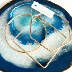 May include: Gold-toned geometric bangles in a circle, a half-circle, and a pentagon shape. The jewelry is displayed on a blue and white agate slice. A white tag with the text "Maimoda Handmade in Hawaii" is attached.
