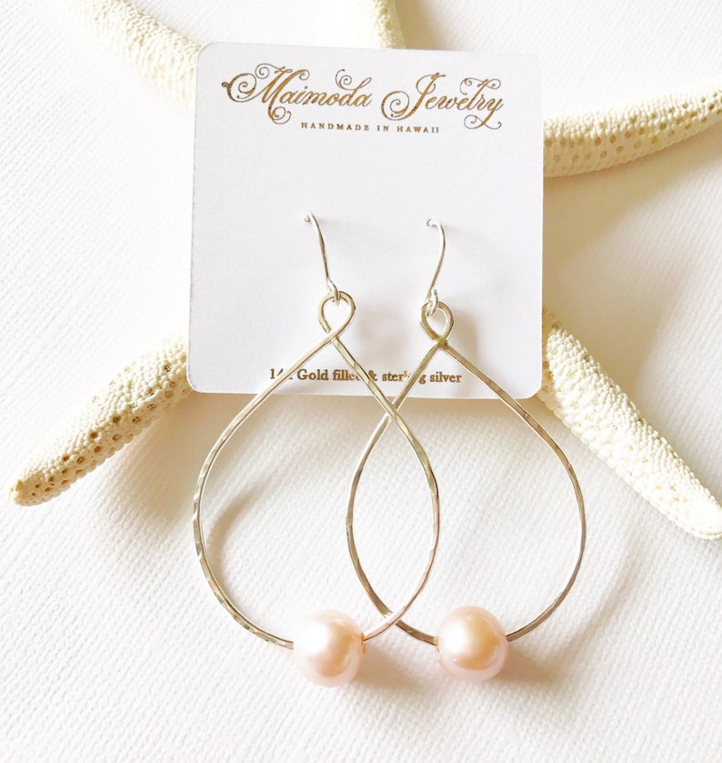 May include: A pair of silver teardrop-shaped earrings with a delicate hammered texture. Each earring features a small pink pearl hanging from the bottom.