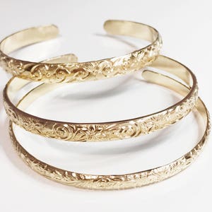 May include: Three gold bracelets with intricate floral designs. The bracelets are stacked on top of each other.