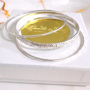 May include: Three silver bangles are displayed on a white surface. Two are plain, while the middle one features a floral pattern. The bangles rest on a gold-colored surface with embossed text. The jewelry is presented in a white box.