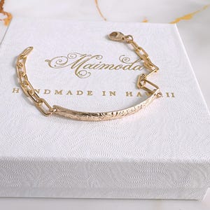 May include: Gold bracelet with a delicate chain and a curved, engraved bar. The bar has a floral design and the text "HANDMADE IN HAWAII".