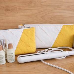 May include: Two white and yellow cosmetic bags with a flat iron, a white mug, and beauty products. The bags have a diagonal yellow design. The beauty products include two bottles of "rosemary mint" products.