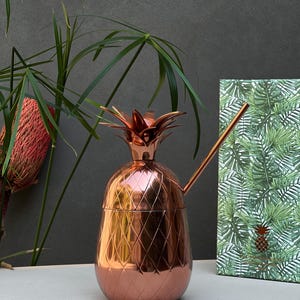 Può includere: Un bicchiere da cocktail in metallo color rame a forma di ananas con coperchio rimovibile dotato di una parte superiore a foglie. Il bicchiere presenta un motivo a rombi testurizzato all'esterno e include una sottile cannuccia in rame.