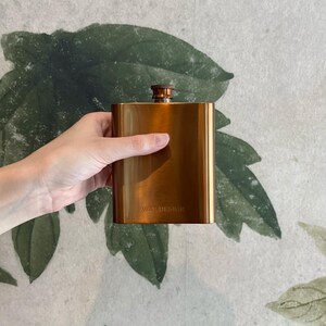 Könnte beinhalten: Ein metallischer kupferfarbener Flachmann mit rechteckigem Körper und abgerundeter Oberseite. Der Flachmann verfügt über einen Schraubverschluss und den Schriftzug W&P DESIGN, der auf der unteren Vorderseite eingeprägt ist. Die Oberfläche ist glatt.