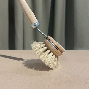 Könnte beinhalten: Eine Spülbürste mit einem langen, hellen Holzgriff und einem runden Holzkopf. Die Bürste verfügt über steife, helle Naturborsten, die mit einem silberfarbenen Metallverbinder am Griff befestigt sind und zum Reinigen von Geschirr dienen.