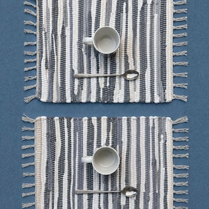 May include: Two rectangular woven placemats featuring a vertical striped pattern in shades of white, light gray, and dark charcoal gray. Each placemat is finished with knotted fringe on the left and right sides and holds a ceramic mug and spoon.