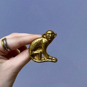 Puede incluir: Una pequeña figura de mono de metal dorado con una superficie texturizada. El mono se muestra en posición sentada con la cabeza girada hacia un lado, mostrando rasgos faciales detallados y una cola larga que se curva a lo largo de la base.
