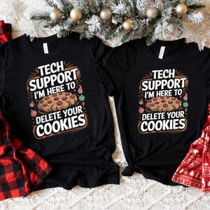 May include: Two black short-sleeve t-shirts featuring a graphic print with the text TECH SUPPORT I'M HERE TO DELETE YOUR COOKIES. The design includes a central illustration of a chocolate chip cookie surrounded by small Christmas ornaments.