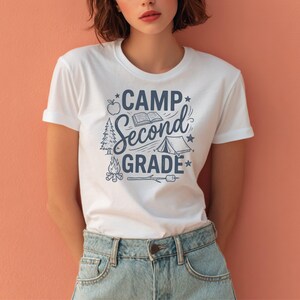 May include: A white short-sleeved t-shirt featuring a dark blue graphic print with the text CAMP SECOND GRADE. The design includes illustrations of an apple, an open book, a pine tree, a tent, a campfire, and a marshmallow on a roasting stick.