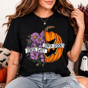 Peut inclure: Un t-shirt noir avec un motif graphique représentant une citrouille coupée en deux. Un côté montre des fleurs violettes, tandis que l'autre est une citrouille d'Halloween orange. Une bannière blanche au centre indique Sorta Sweet et Sorta Spooky.