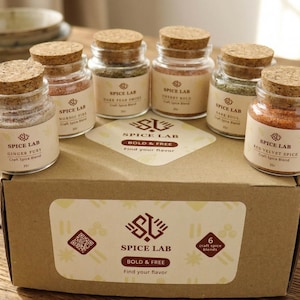 Könnte beinhalten: Sechs Gewürzmischungen in kleinen Gläsern mit Korkdeckeln, präsentiert auf einer Pappschachtel. Die Gläser sind mit dem "Spice Lab"-Logo und verschiedenen Gewürznamen beschriftet. Die Schachtel zeigt auch das "Spice Lab"-Logo und den Text "Bold & Free".