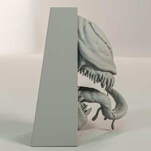 Pu&ograve; includere: Una scultura grigia stilizzata di un cranio e di una mascella di creatura, con denti affilati e una lingua curva. La scultura &egrave; parzialmente nascosta da un supporto rettangolare grigio.