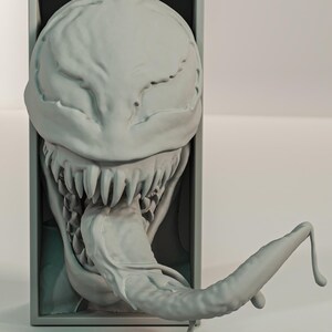 Pu&ograve; includere: Una scultura grigia stampata in 3D di una testa mostruosa con denti affilati e una lunga lingua estesa. La testa &egrave; inserita in una cornice rettangolare e il design generale ha un aspetto strutturato e organico.