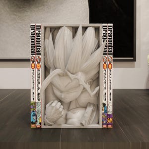 Puede incluir: Escultura blanca de un personaje de Dragonball Z con pelo puntiagudo, enmarcada por dos vol&uacute;menes del manga Dragonball. Las lomos del manga son visibles, con texto japon&eacute;s e ilustraciones coloridas. La escultura est&aacute; sobre una superficie oscura.