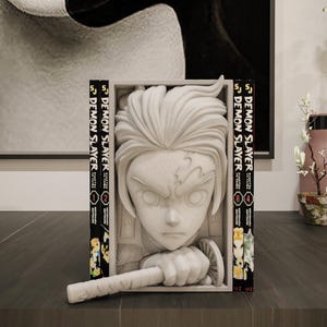 Könnte beinhalten: Eine weiße Skulptur einer Figur aus der Anime-Serie Demon Slayer, eingerahmt von zwei Manga-Bänden. Die Skulptur steht auf einer dunklen Oberfläche, mit einem verschwommenen Hintergrund mit Kunstwerken und dekorativen Gegenständen.