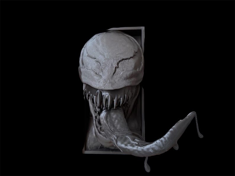 Pu&ograve; includere: Scultura grigia tridimensionale di una testa di creatura con una grande bocca, denti affilati e una lunga lingua. L'opera &egrave; posta su sfondo nero, evidenziando i dettagli e la consistenza.