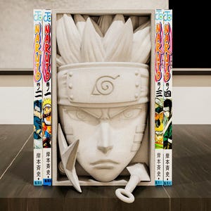 May include: A white sculpture of a character's face with spiky hair, a headband, and a stern expression. Two white kunai and a ring-shaped weapon are in front of the sculpture. Two manga books with colorful covers are on either side of the sculpture.