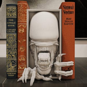 Pode incluir: Uma escultura alienígena branca com dentes e garras afiadas está posicionada entre três livros. Os livros são intitulados "The Jungle Book" e "Virginia's Venture". Os livros são em tons de azul e laranja.