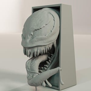 Pu&ograve; includere: Una scultura grigia stampata in 3D di una testa di mostro che emerge da una cornice rettangolare. Il mostro ha denti affilati, una lunga lingua e una superficie strutturata. La scultura &egrave; un'opera d'arte unica.