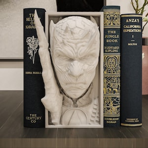 May include: A white sculpture of a face with a crown and a sword, used as a bookend, with three dark blue books. The books have gold lettering and the titles include "The Jungle Book" and "Anza's California Expeditions".