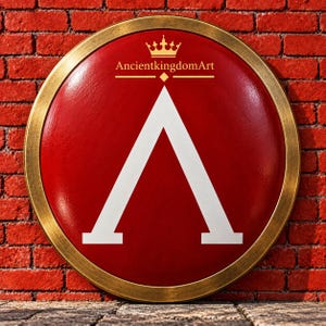 Op de afbeelding: Een rond schild met een dieprood oppervlak en een grote witte Griekse letter Lambda in het midden. Het schild heeft een metalen goudkleurige rand en toont de tekst AncientkingdomArt in goud onder een klein kroonpictogram bovenaan.
