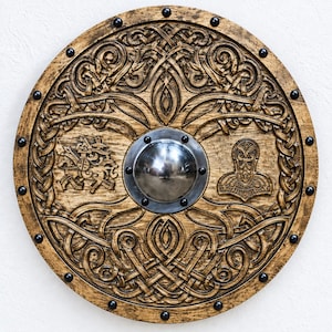 May include: A circular wooden shield featuring intricate carved knotwork patterns, a central polished metal dome, and dark metal rivets along the outer edge. The wood has a distressed brown finish with carved symbols on the left and right sides.